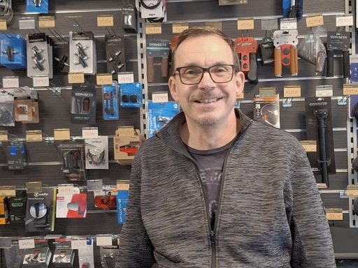 Ladeninhaber Stefan Stark steht vor einer Wand mit Ersatzteilen. Er trägt eine graue Jacke und eine Brille.