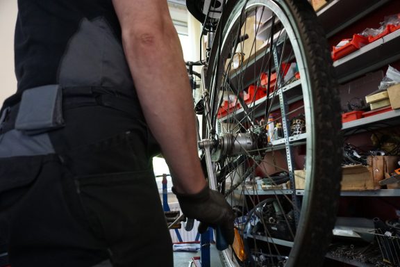 Ein Mann in Arbeitskleidung repariert das Hinterrad eines Fahrrades. 